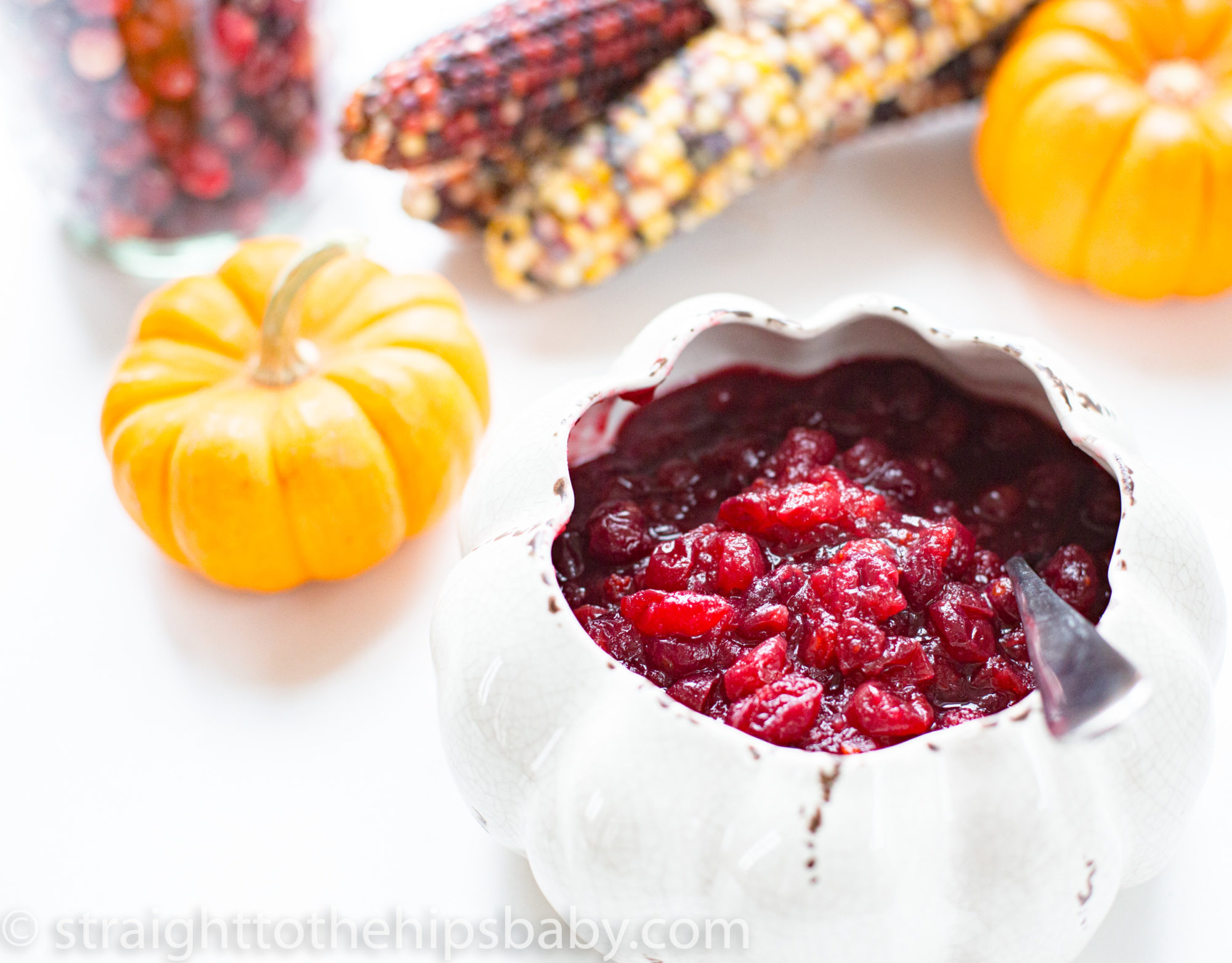 A white pumpkin-shaped bowl filled with cranberry sauce, alongside a small orange pumpkin and colorful dried corn. The vibrant, chunky cranberry sauce contrasts against the light background, evoking a festive autumn or Thanksgiving setting.