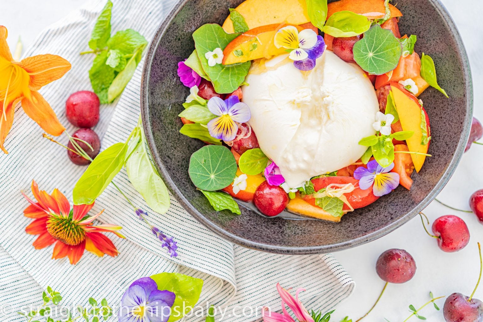 A bowl of fresh salad with a ball of burrata cheese, surrounded by vibrant fruits and edible flowers. The salad includes peach slices, tomatoes, and cherries, garnished with green leaves. Edible flowers and cherries are also scattered around the bowl on a striped cloth.